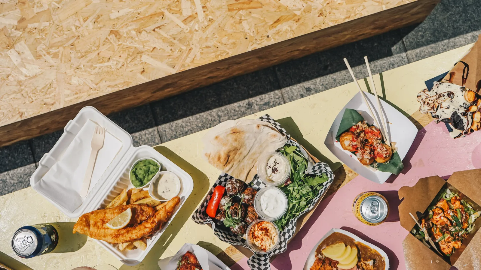 Overhead view of takeout food on an outdoor table: fish and chips in a box, a tray of flatbread, salad and dips, a curry, and canned drinks