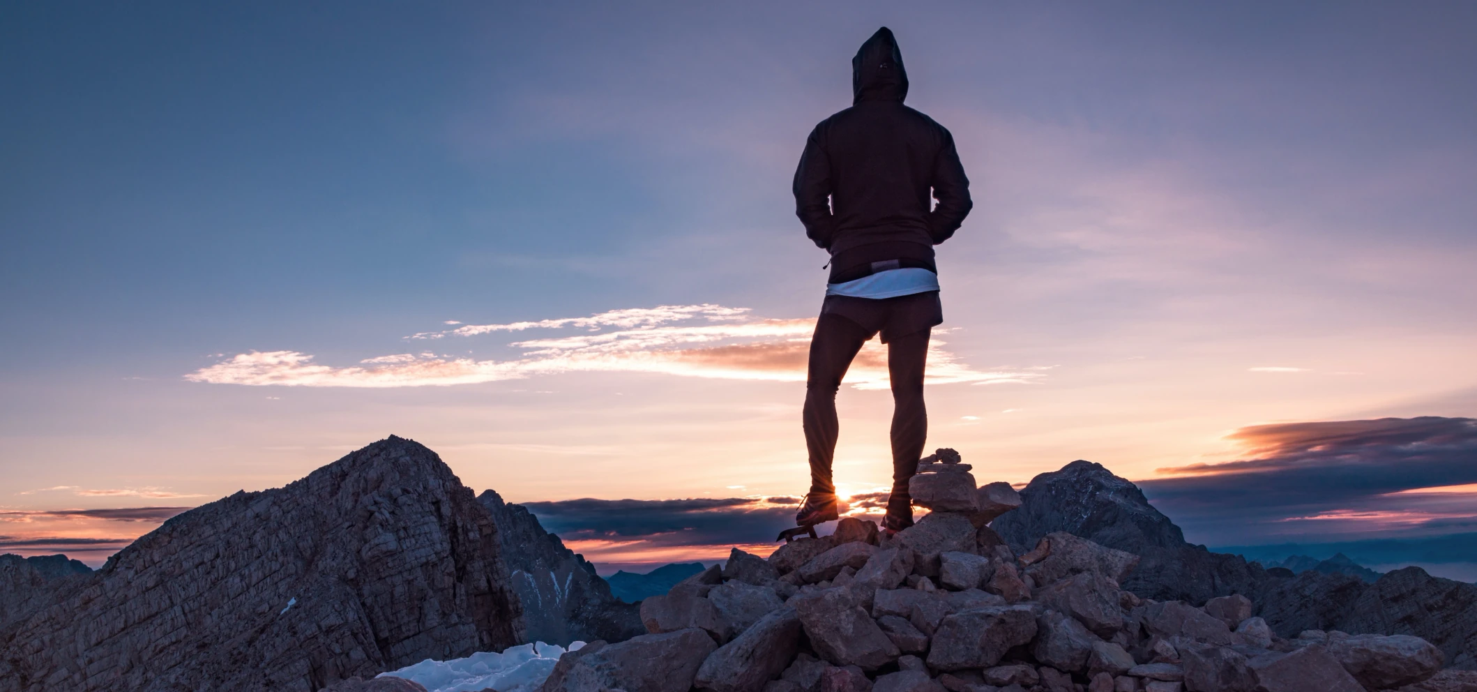 A person in a hooded jacket standing on a rocky mountain summit, seen from behind, facing a sunset sky