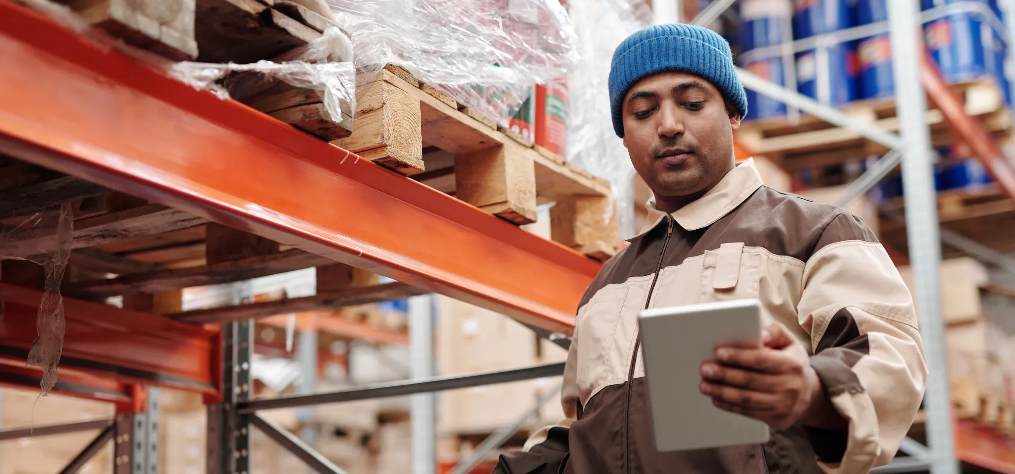 A warehouse worker in a blue knit cap reading a tablet beside orange pallet racks