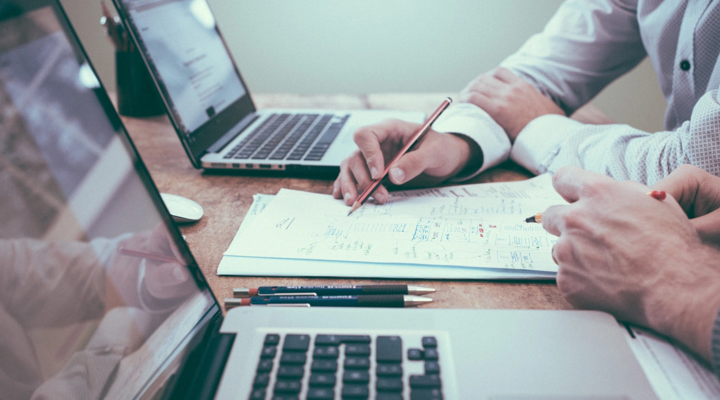 Two people at a desk with laptops, one marking up printed notes with a pencil